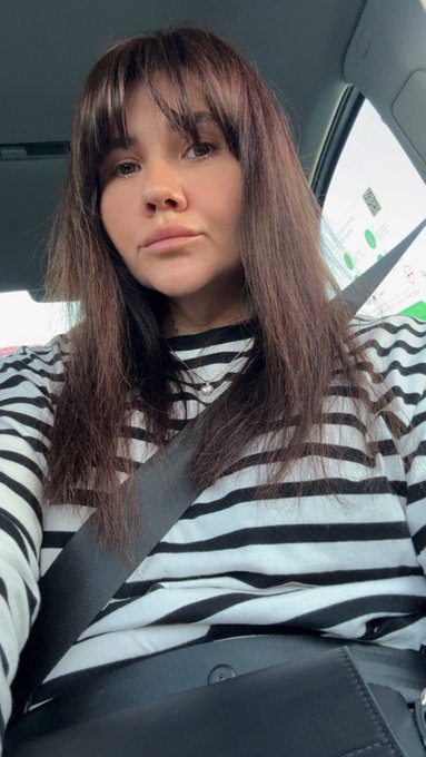 A selfie of a young woman with long dark hair and bangs, wearing a black and white striped shirt and a seatbelt while sitting in a car.