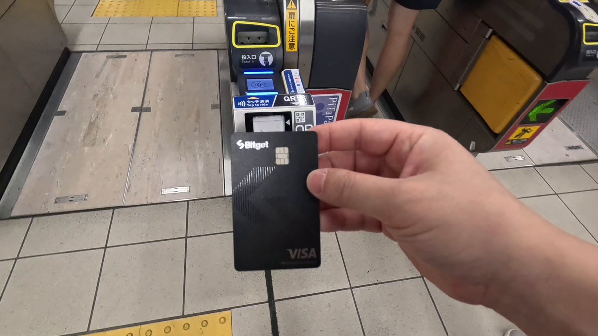 The image shows a person holding a black Visa card with a Visa logo on it. The person is standing next to a subway station, possibly waiting for their train or just leaving after using public transportation. The focus of the image is on the person's hand holding the credit card and not on any other objects in the scene.