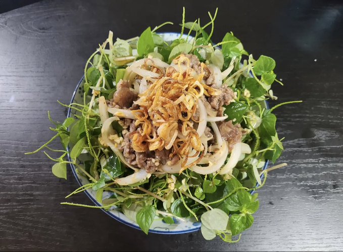 A Vietnamese-style salad on a blue-rimmed plate featuring beef, onions, fried shallots, fresh herbs, and peppery greens on a dark wooden surface.