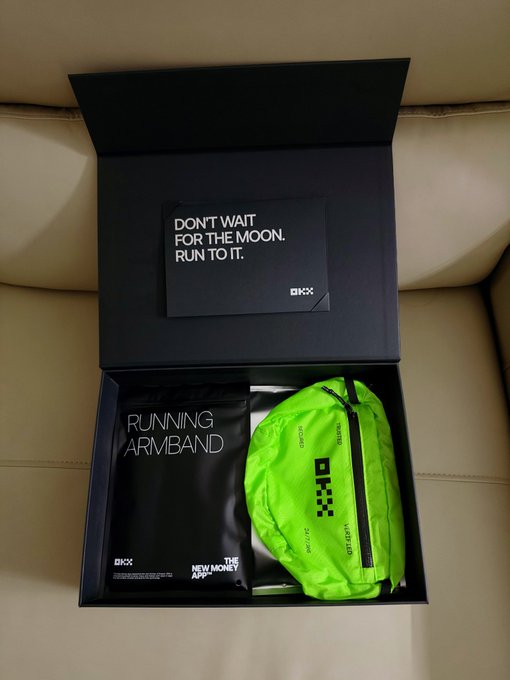 An open box on a tan couch containing a black 'RUNNING ARMBAND' package and a bright neon green running belt, with a card reading 'DON'T WAIT FOR THE MOON. RUN TO IT.'