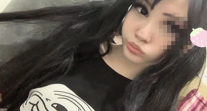 A selfie of a young woman with long dark hair, wearing a black shirt with a cartoon graphic, blowing a kiss at the camera with a pixelated area obscuring part of the image.