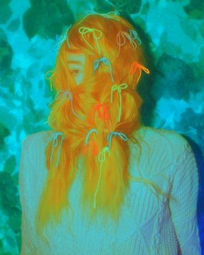An inverted-color photograph showing a person with long hair adorned with small glowing string lights, wearing a textured sweater against a leafy background.