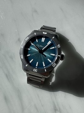 A silver wristwatch with a teal textured dial resting on a white marble surface, casting a shadow.
