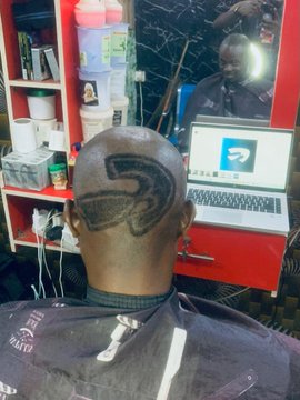 A man sitting in a barber chair getting a large 'S' shaved into the back of his head, with a laptop visible in the mirror's reflection.