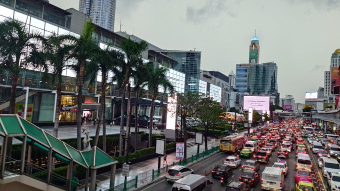 An urban cityscape showing a busy street with heavy traffic congestion, modern glass buildings, palm trees, and skyscrapers in the background.