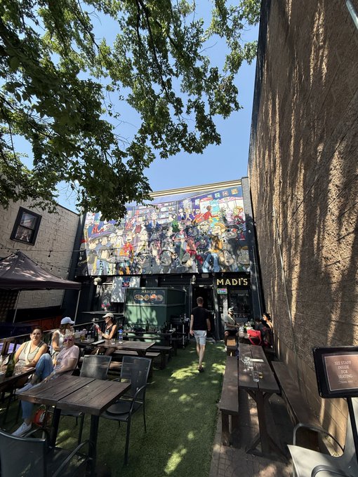 An outdoor patio area of a restaurant called 'MADi's' with people seated at tables on artificial turf, with a large colorful mural on the back wall and a sign reading 'SORRY OUTSIDE SEATING ONLY'.