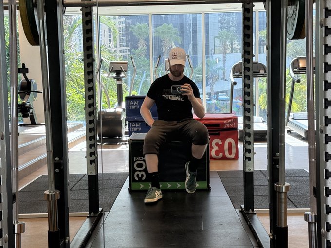 A man in a black t-shirt and cap sits inside a squat rack at a gym, posing for a photo with a box labeled "30 INCH" visible behind him. The image supports the post's message about strength standards in the @monad crypto community, suggesting that members should prioritize physical fitness benchmarks like squatting 315 pounds before complaining about difficulty breaking into the group.