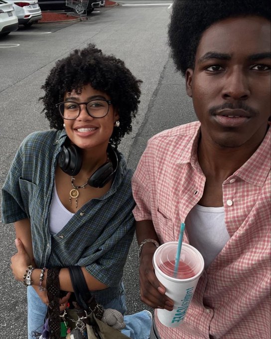 A casual selfie-style photo of a smiling young woman with curly hair and headphones around her neck standing next to a young man holding a drink in a parking lot.