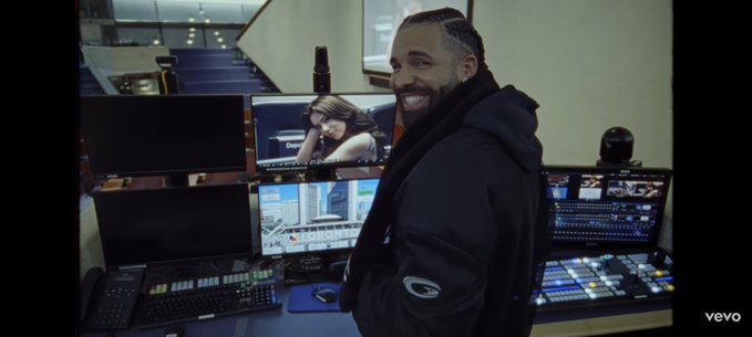 A smiling man with braided hair, resembling Drake, stands in a production control room with multiple monitors displaying various video feeds.