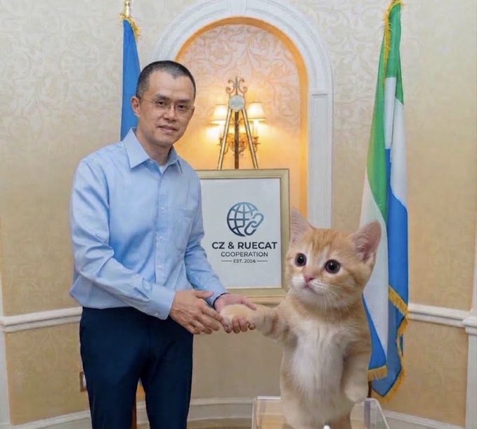 A man (CZ, Changpeng Zhao) shaking hands with a ginger kitten in front of a 'CZ & RUECAT COOPERATION' sign and Binance flags.