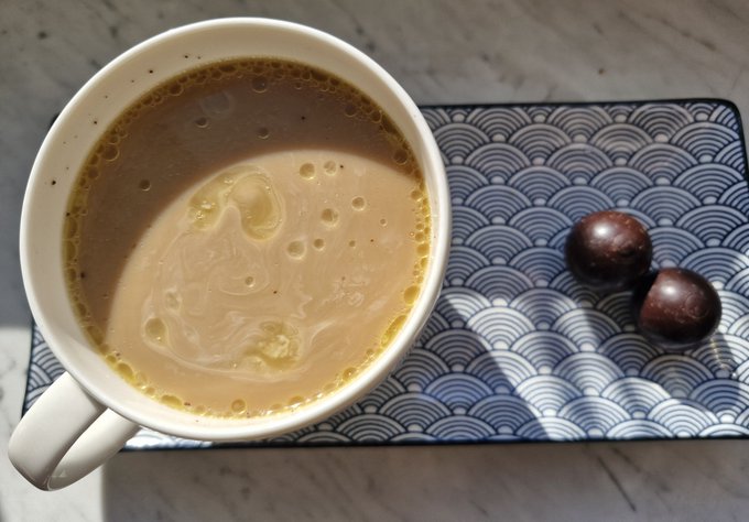 A white mug of milky coffee or tea with foam bubbles sits on a blue and white patterned plate, accompanied by two dark chocolate candies.