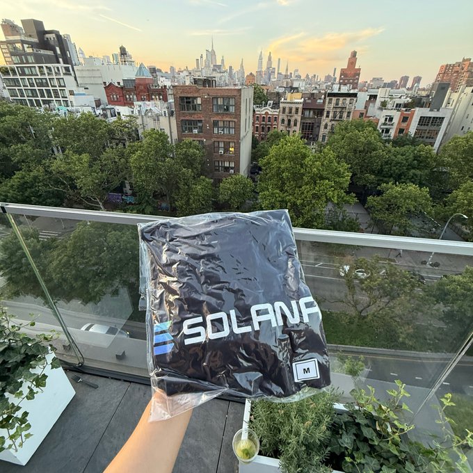 The image shows a person holding up a black shirt with the word "Solana" written on it, which appears to be a new purchase or gift for someone else. The person's hand is visible above the shirt, while the rest of their body is not shown in the photo. The background features a cityscape and trees, suggesting that this scene takes place in an urban setting with greenery nearby.