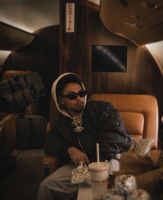 A stylish portrait of a young man wearing sunglasses, a hoodie, and a large pendant necklace, seated inside a dimly-lit private jet or luxury lounge.