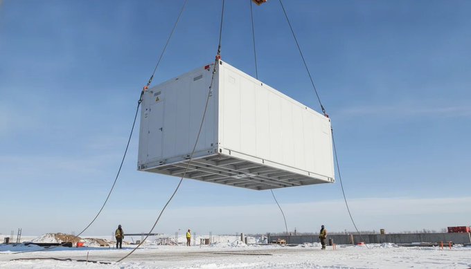 A large white shipping container—likely housing AI data center infrastructure—is suspended by crane cables in a snowy outdoor site, with workers in safety gear visible below. The image visually illustrates Empery Digital's $65 million AI data center project, partially funded by its recent sale of 1,400 Bitcoin.