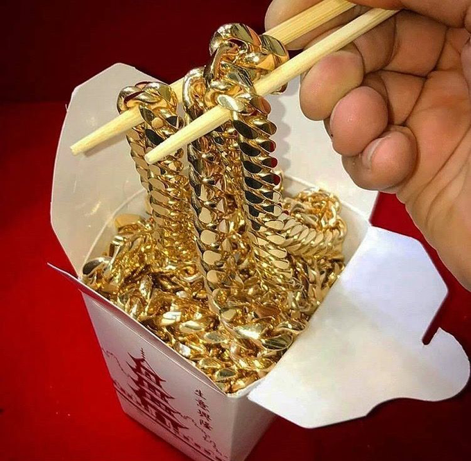 A hand uses wooden chopsticks to lift shiny, gold-colored chain necklaces from a white Chinese takeout box against a red background, with a pagoda illustration and Chinese characters visible on the side.