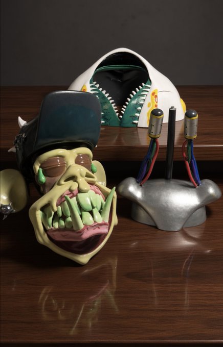 A collection of artistic objects sits on a wooden surface, including a sculpted head wearing a green baseball cap, a green-toothed mouth mask, and a silver base with attached wires and cylindrical components.
