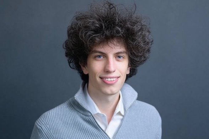 A young man with voluminous curly brown hair smiles warmly at the camera, wearing a gray knit sweater over a white collared shirt against a gray backdrop. The image humorously references "Sam Bankman-Aschenbrenner," with the man's wild, untamed hairstyle humorously playing on the "Aschenbrenner" (German for "ash burner") pun, suggesting his head is literally on fire.
