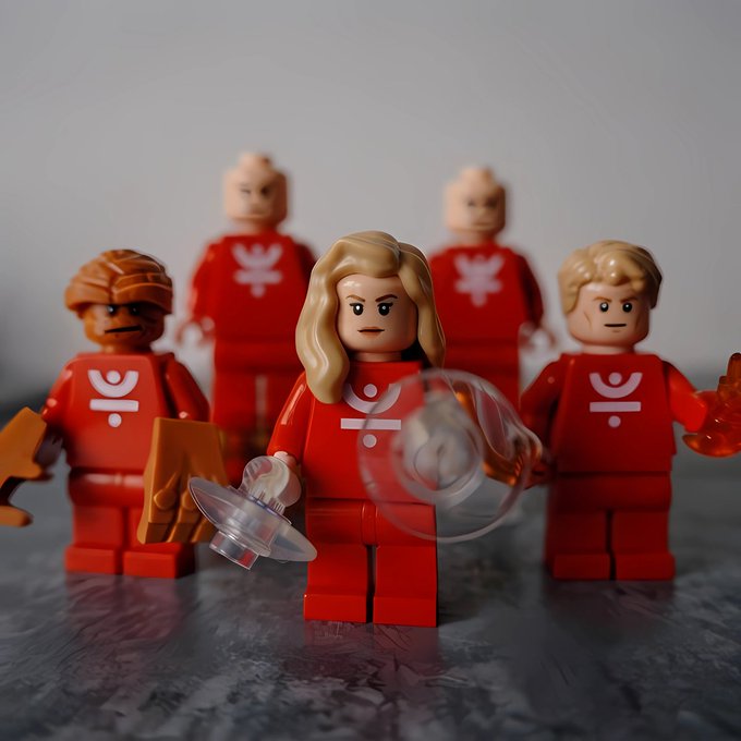 A photograph of LEGO minifigures dressed in red uniforms with a logo resembling a person, featuring a central blonde female figure holding a swirling energy effect.
