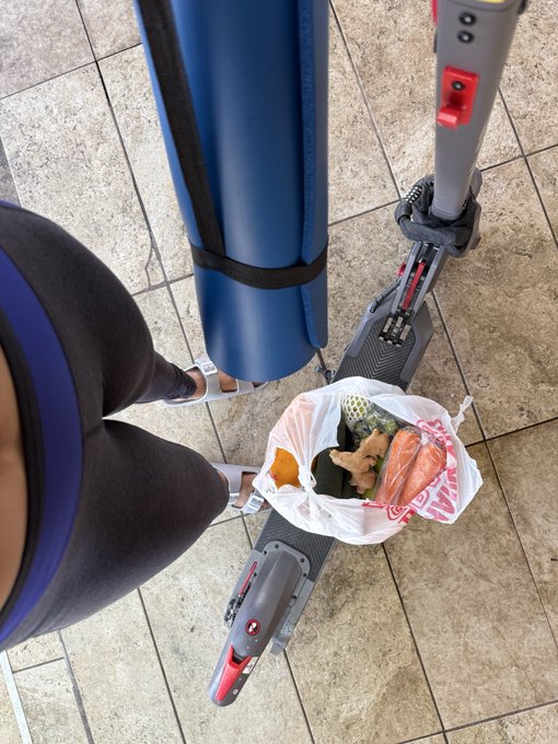 A first-person perspective photo looking down at a person's legs in dark athletic shorts and sneakers, standing next to an electric scooter that has a plastic bag of groceries hanging from the handlebars.