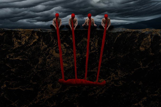 A surreal, dark art image showing four shirtless figures with red hoods sitting on a cliff edge, each connected by a long red tube to a red form lying at the bottom of a rocky pit.