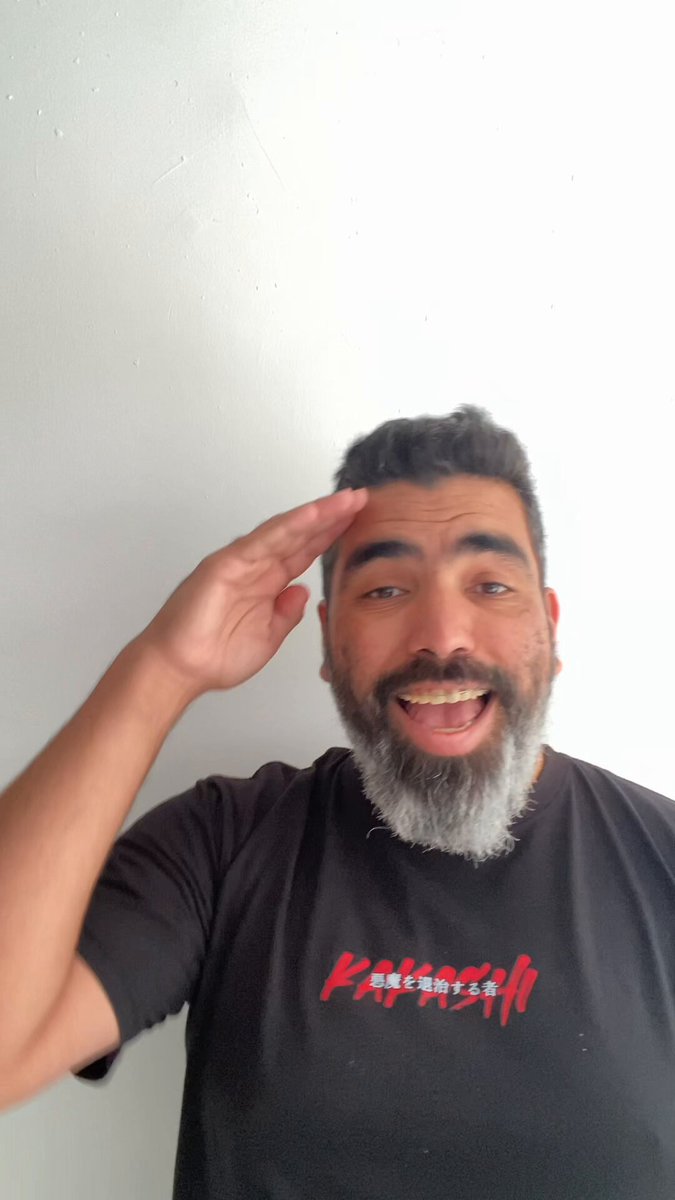 The image features a man with a beard wearing a black t-shirt that says "Kiss the gunk" or "@BagsHackathon". He has his hand raised, possibly saluting or showing appreciation for something. The background is white, which contrasts with the black t-shirt and makes it stand out. There are also some text elements in the image, such as "@solana", "@SP3NDdotshop", and "Thank you so much, @BagsHackathon I still can't believe it." These details provide context to the man's outfit and the post he is sharing on social media.