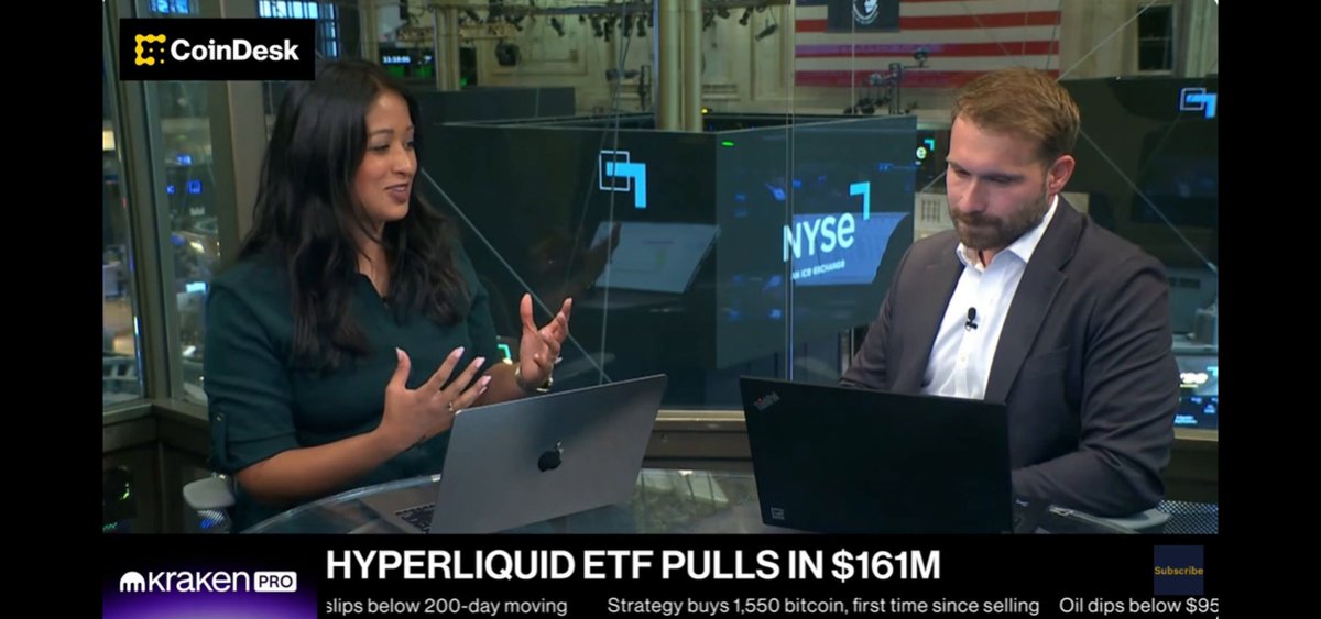 The image shows a news segment featuring two people, one woman and one man, sitting at a table with their laptops open. They appear to be discussing something related to cryptocurrency or the stock market. The background of the scene includes multiple monitors displaying various financial data, indicating that they are likely analyzing market trends and making investment decisions.