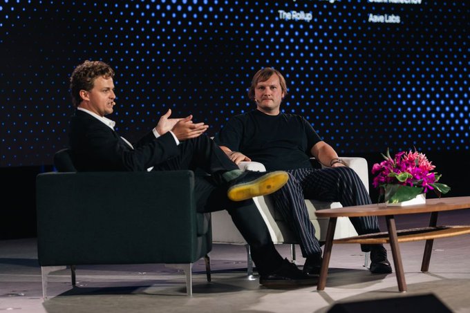 Two men sit on stage during a panel discussion, with one gesturing while speaking to the other, against a backdrop featuring "Aave Labs" branding alongside text reading "The Pickup" and "Interop." The image complements the post by visually capturing the conversation with Stani Kulechov, CEO of Aave Labs, at the TOKEN2049 Singapore event.