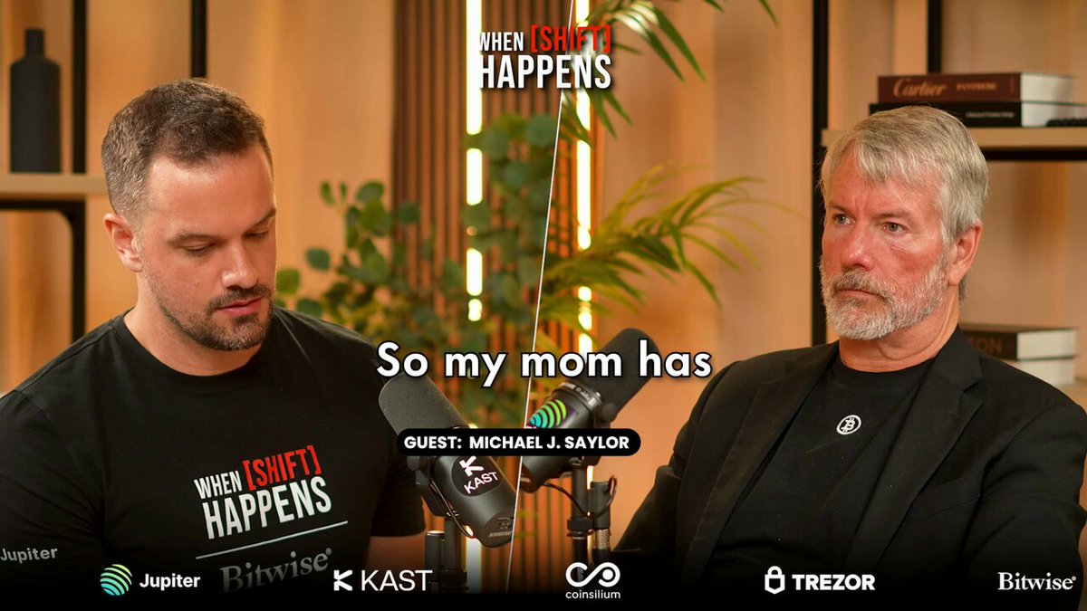 The image features two men wearing black t-shirts, one of which says "So my mom has" while the other reads "Strategy". They are sitting next to each other with a microphone in front of them, suggesting they might be discussing or interviewing someone related to Bitcoin.