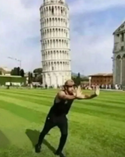 Video still showing a muscular man in a black tank top posing in front of the Leaning Tower of Pisa on a sunny day with green grass.