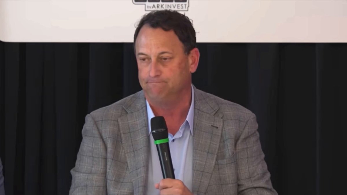 The image shows a man wearing a grey suit jacket holding a microphone at a conference or event. The man appears to be speaking into the microphone while standing behind a podium. The background of the image features a white wall with black text, which includes the words "Palantir" and "ARKInvest".
