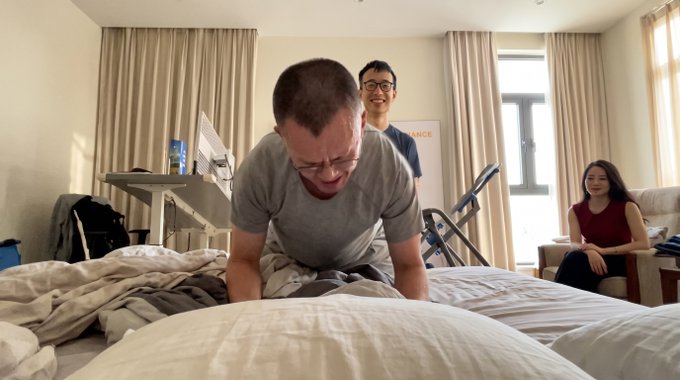 A man in a gray t-shirt sits on a bed laughing, with another smiling man standing behind him and a woman seated on a couch in a room featuring an exercise bike and a "BINANCE" banner. The "one chair" caption humorously highlights the irony in the image: despite the room containing multiple seating areas—a chair, couch, and bed—everyone ends up lounging wherever is convenient, much like the unpredictable, "sit wherever" nature of trading on Hyperliquid.
