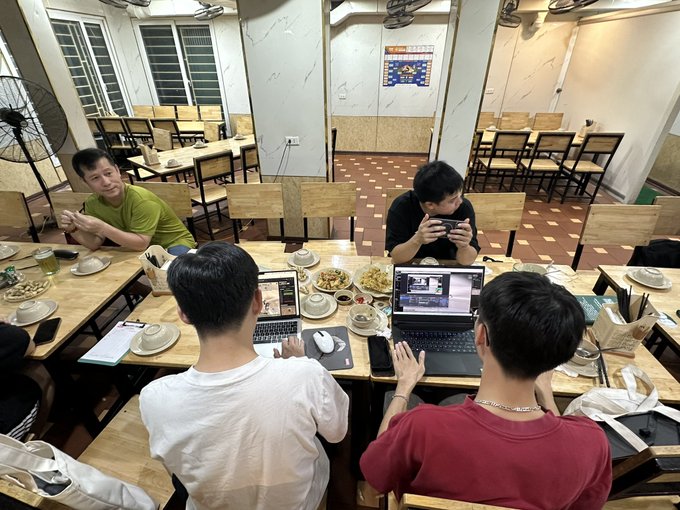 A group of four young men gathered around a table in a restaurant, with two using computers/laptops and one looking at a phone, surrounded by food and drink.
