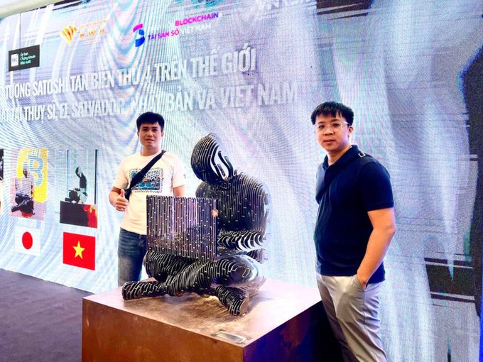 Two men posing beside a black humanoid sculpture at what appears to be a SatoShin Blockchain art exhibition in Vietnam.