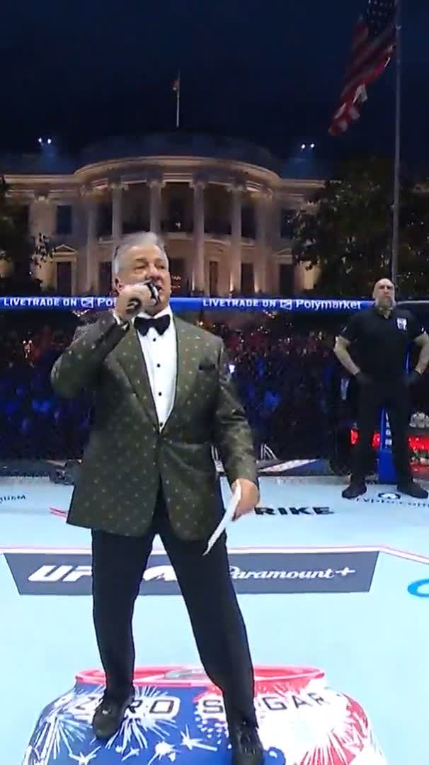 The image features a man dressed in a suit, standing on top of a large soda can that has been wrapped with foil paper. The man appears to be singing into a microphone while wearing a bow tie. In the background, there are two men present - one is holding a flag and another person is standing behind him. The backdrop includes an American flag, which suggests that this event might be taking place in front of the White House or a similar significant location.