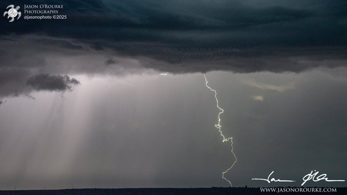 A dramatic photograph by Jason O'Rourke (@jasonophoto ©2025) captures a lightning bolt striking down from a dark, stormy sky with sun rays piercing through the clouds over a flat horizon.