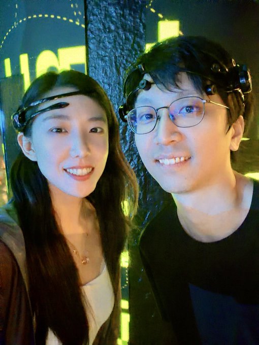The image features a man and woman standing next to each other, both wearing glasses with headbands on their heads. They are smiling for a photo, possibly at an event or gathering where they have been given special headbands as part of a challenge or game. The background is dark, which contrasts with the bright smiles on their faces. The image also includes text that reads "vibe coding to code".