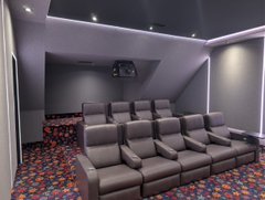The image shows a large, empty room with gray walls and a purple ceiling. There are multiple rows of brown leather chairs arranged facing each other, creating an impression of a theater or auditorium setting. The chairs appear to be well-lit by the purple lighting system installed in the ceiling. A television is mounted on the wall above the seating area, suggesting that this room could serve as a space for watching movies or performances.