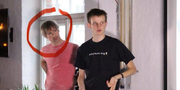 Two men standing indoors, with the man in front wearing a black shirt with 'ethereum.org' printed on it, while the man behind in a red shirt is circled in red marker.