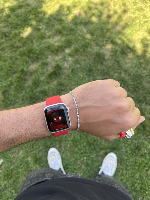 A person shows off their Fitbit Versa with a red band featuring a Sith/Darth Vader watch face, paired with a diamond tennis bracelet, while standing on grass in white sneakers—perfectly capturing the "touching grass" and "locked in" vibe of the post.