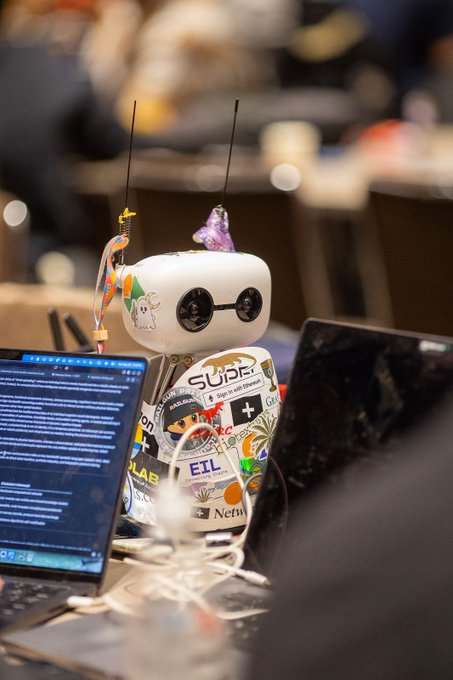 A small decorative robot covered in stickers, including 'SUPE!', 'EIL', and 'Nex', sits on a desk between a laptop displaying code and another laptop, adorned with a tiny party hat and antennae.