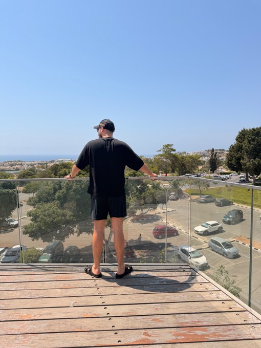 A man in a black t-shirt, shorts, and cap stands on a wooden deck overlooking a coastal town and parking lot, soaking in the morning view. The serene seaside scenery pairs perfectly with a cheerful "Gm friends" greeting, capturing a peaceful start to the day.