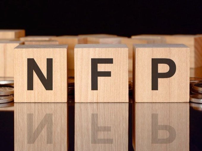 Three wooden blocks displaying the letters "NFP" sit atop a reflective surface, with stacks of coins visible in the background. The image directly illustrates the post's reference to the Non-Farm Payrolls (NFP) report, a key economic data release that often triggers volatility in markets including crypto.