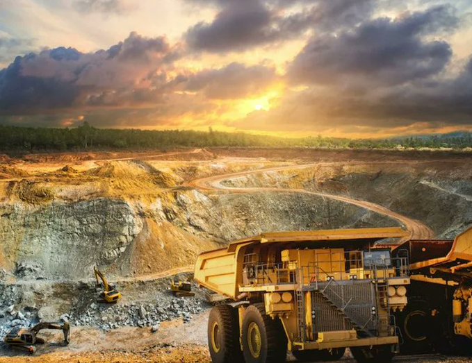 A landscape photo of a large open-pit mine at sunset, featuring a massive yellow haul truck in the lower right and smaller excavators working in the rocky terrain.