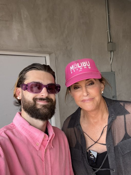 A selfie photo of a bearded man wearing purple sunglasses and a pink shirt, posing next to a woman wearing a black sheer top and a bright pink 'Malibu Strong' trucker hat