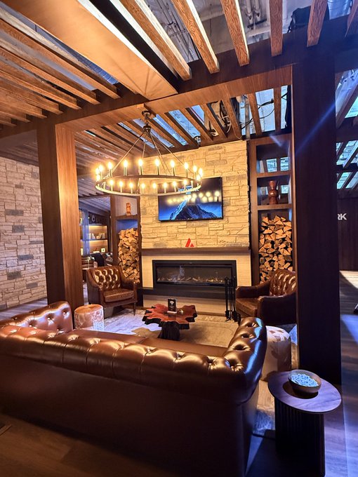 A warmly lit, rustic-style lounge or lobby featuring brown leather sofas, a fireplace, wooden beamed ceiling, stacked firewood, and a circular chandelier.