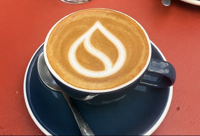 A dark blue ceramic cup of coffee featuring a leaf latte art design, served on a matching saucer with a spoon on a red table.