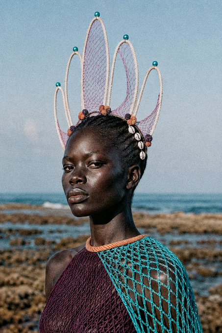 A model with braided hair wears an elaborate pink crown-like headpiece adorned with cowrie shells, standing near the ocean in a teal mesh garment.