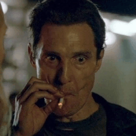 A man with a gaunt, intense expression smokes a cigarette in a dimly lit setting, holding it close to his face.