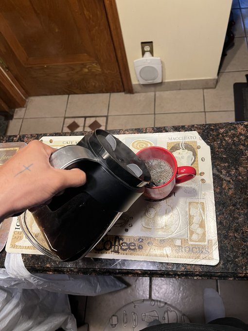 Photo of a hand with a cross tattoo pouring hot water from a black electric kettle into a red mug on a kitchen counter with a coffee-themed placemat.