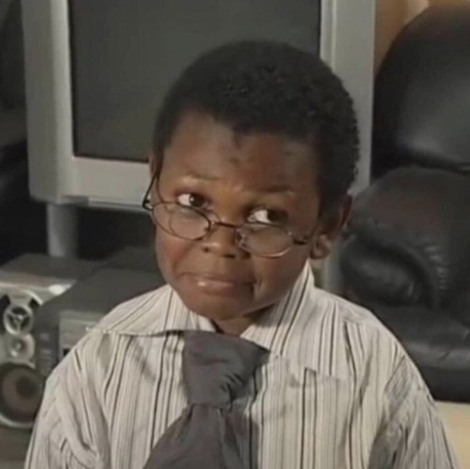 A young boy wearing glasses, a striped button-up shirt, and a loose tie makes a skeptical or unimpressed facial expression while seated in front of an older computer monitor.